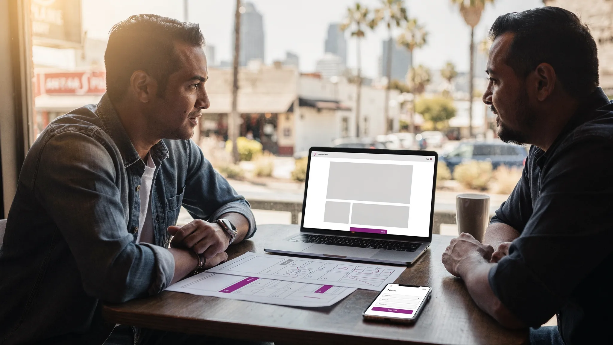 A Los Angeles small business owner meeting with a local web developer at a table with printed website wireframes, a laptop (screen facing the viewer but showing a neutral blank layout), and a phone with a simple contact form mockup. In the background, a subtle LA cityscape and storefront details suggest a local setting.