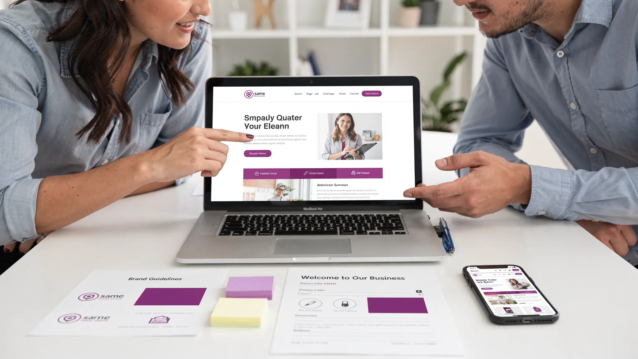 A small business owner and a web designer reviewing a website homepage mockup together on a laptop at a desk, with printed brand notes and a phone showing a live preview of the mobile layout.